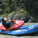 2 people in blue helmets with a red kayak on top of a blue inflatable raft in a river.