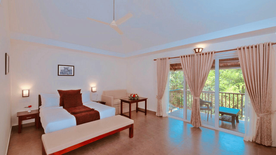  A neatly arranged room with twin beds, a wooden bench, and large glass doors opening to a balcony with scenic views.- Abad Brookside Lakkidi, Wayanad