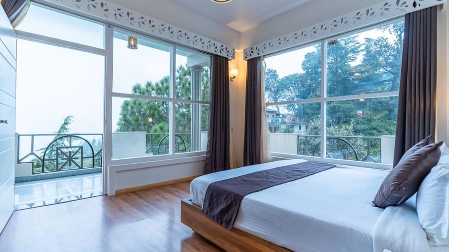A bedroom with a white bed, a rug, and large windows, interior setting - Clarks Exotica Resort, Near Mukhteshwar.
