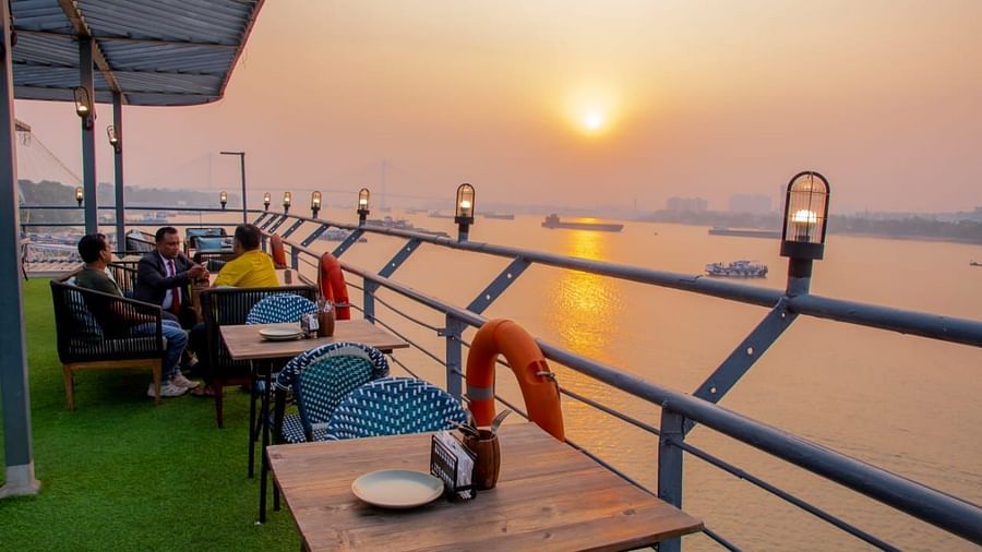 People sitting at tables with a sunset view over a river, seen from a rooftop bar with a grassy floor and metal railing.