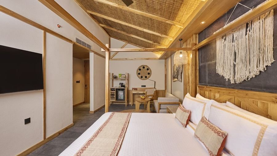 A king size bed with a runner, a TV, a desk, and a minibar in a room with a sloped wooden ceiling at Royal Tulip, Chitwan.