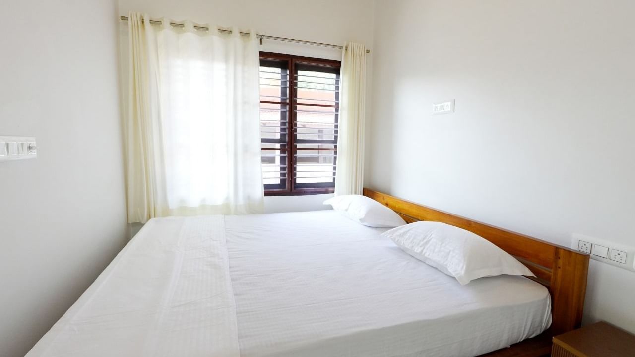 A bright and inviting deluxe room at Bhuthanakadu Retreat, showcasing a comfortable bed with white bedding, a wooden frame, and large windows draped with sheer white curtains.