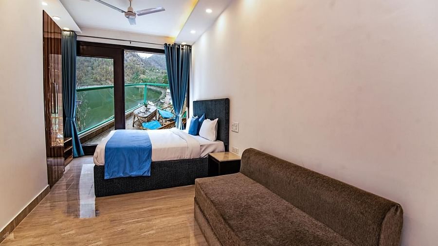 A bedroom with a large bed, sofa, wooden flooring, and full-length windows overlooking greenery.