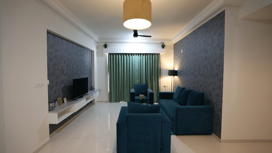 A spacious lounge area with teal armchairs, a wall-mounted television, and floor-to-ceiling green curtains in a luxury apartment at Apartel by Aarin, Oragadam.