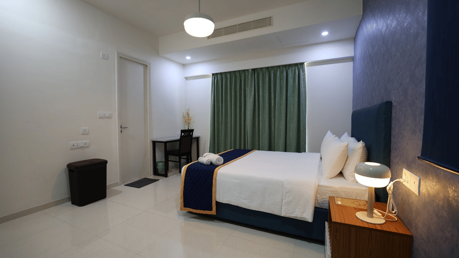 A contemporary hotel bedroom featuring a double bed with a blue runner, a bedside lamp, and a minimalist work desk by the window at Apartel by Aarin, Oragadam.