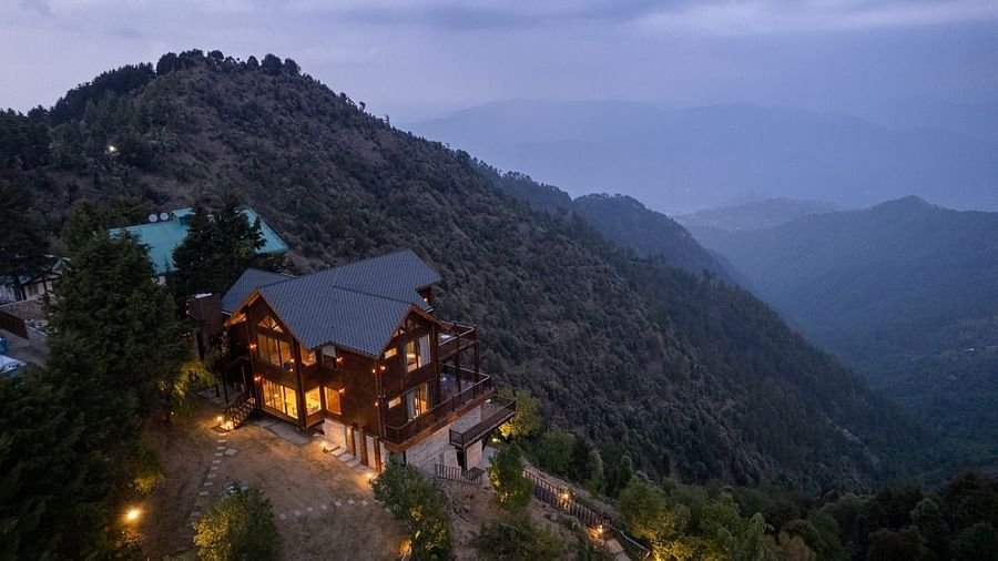 An aerial view of a well lit facade amidst the hilly areas covered with trees and clouds - Avalon Cottages, Kanatal