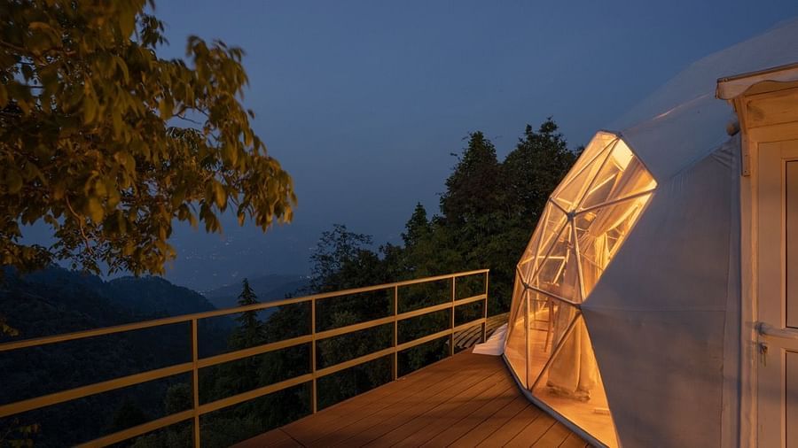 An image of wooden filled with warm lights emitting from a dome style cottage with the backdrop of evening sky filled with clouds -   Avalon Cottages, Kanatal
