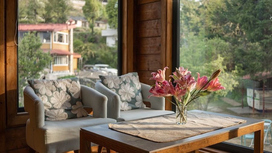 An image of sofa chairs with table near a glass wall from where sunlight is flooding the room | Avalon Cottages, Kanatal