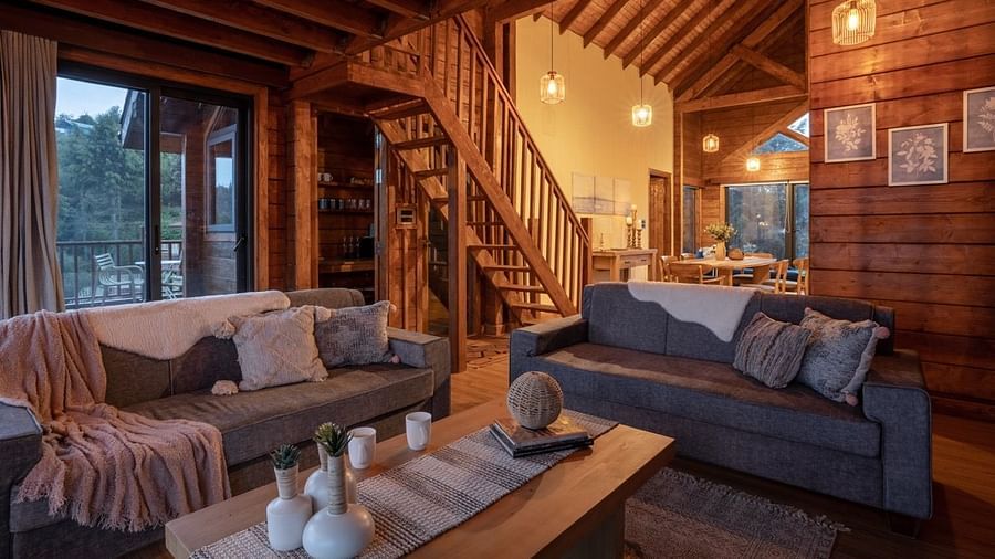 An image of a seating area with sofa set, centre table, frames hanged on wooden walls, and an indoor stair leading to next floor | Avalon Cottages, Kanatal