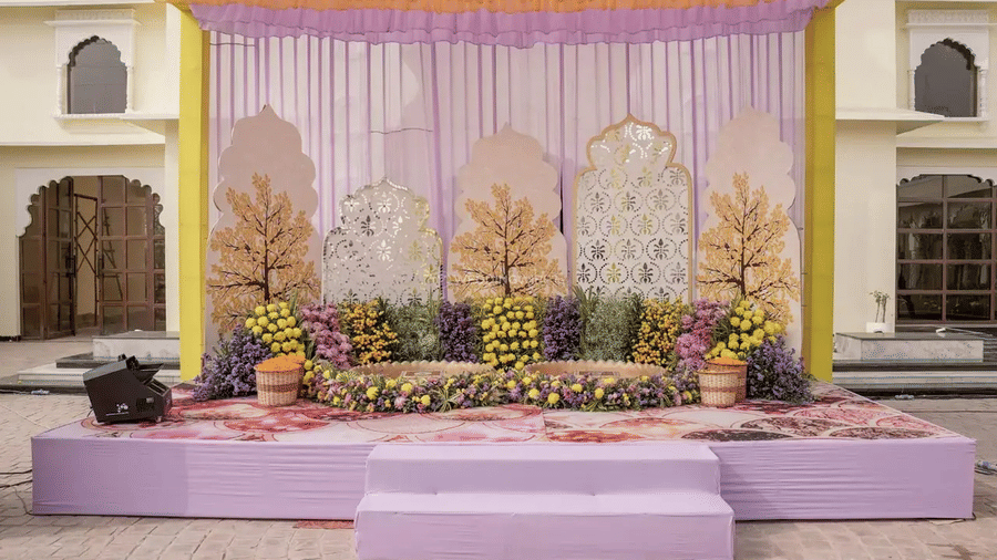 A decorated stage with floral arrangements and tree cut-outs against a purple backdrop at Beelwa Palace, Jaipur
