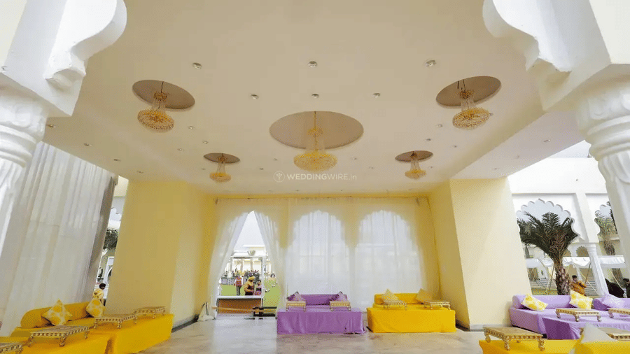 An elegant indoor hall with decorative ceiling, long seating arrangements, and tables at Beelwa Palace, a refined hotel with swimming pool in Jaipur.