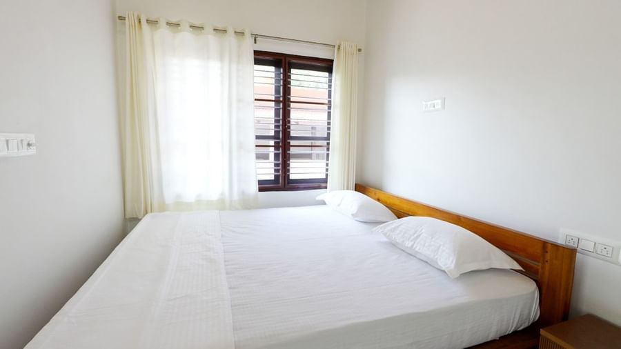 A bright and inviting deluxe room at Bhuthanakadu Retreat, showcasing a comfortable bed with white bedding, a wooden frame, and large windows draped with sheer white curtains.