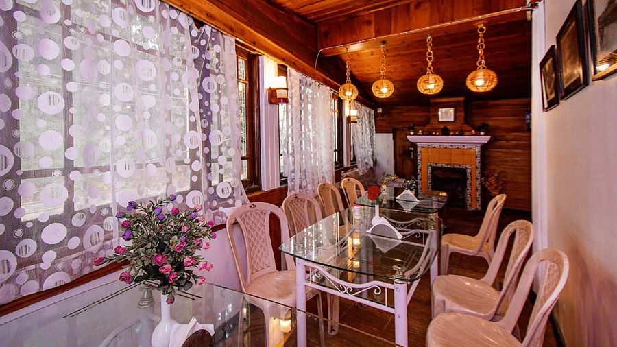 Dining area with a wooden ceiling, hanging lights, a glass table, and floral patterned white lace curtains at Hotel Dolphin Darjeeling.