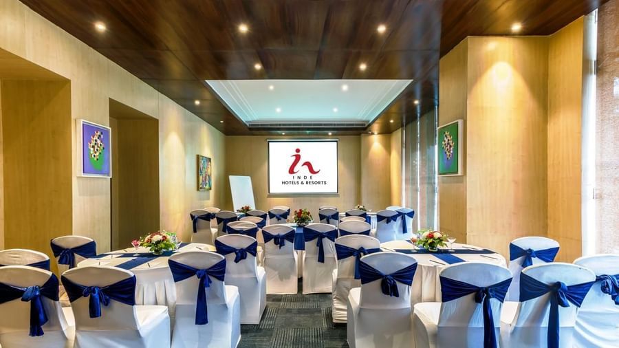 Meeting room at Inde Reva Resorts Rishhikesh, with white chairs tied with navy blue sashes, round tables decorated with flower arrangements, and a projector screen at the front.