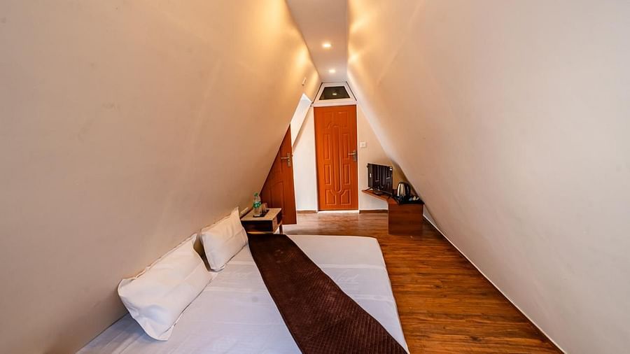 A compact hotel room interior featuring a bed with a dark brown runner, angled light-colored walls, and a small desk area.