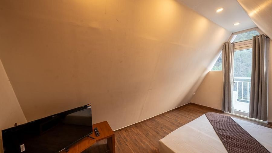 A modern hotel room with sloped light-coloured walls, a bed, a television on a stand, and a large window to the right.
