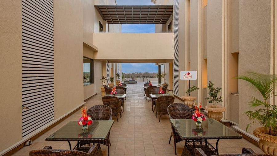 A well-lit outdoor seating area with multiple tables and chairs arranged along a pathway, featuring potted plants and a glimpse of the sky in the background, suggesting a relaxed dining space at Lords Inn Motikhavdi, Jamnagar.