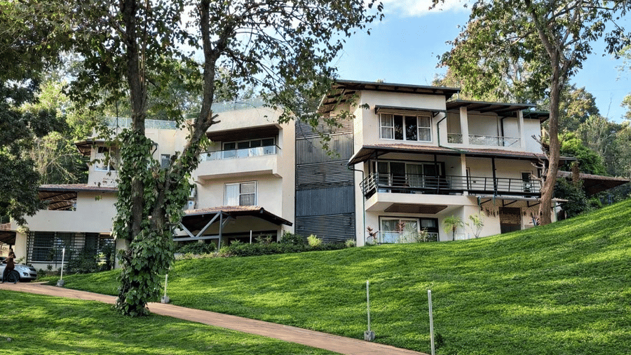 A scenic view of the Lords Riverside Resort, Muduba - Shivamogga building nestled behind a lush green sloping lawn and trees.
