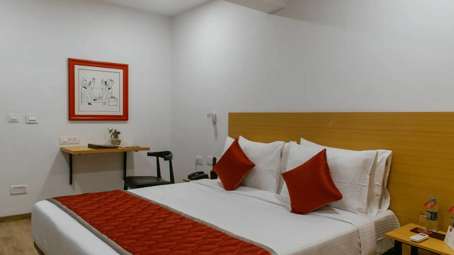 A hotel room featuring a bed with cushions, a framed picture on the wall, a small desk with a chair, and water bottles on the side table - Lords Eco Inn Belagavi