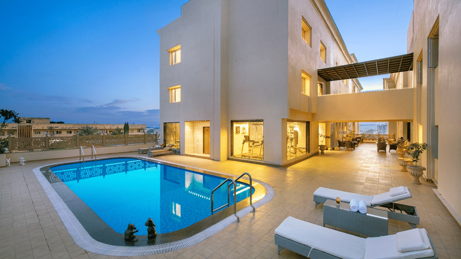 A tranquil outdoor swimming pool area at dusk, with the still blue water reflecting the surrounding light-coloured hotel building, alongside comfortable white sun loungers on the paved area  in Lords Inn Jamnagar.