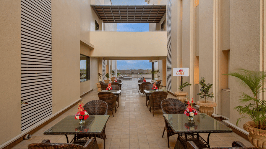 A well-lit outdoor dining area with multiple tables and chairs arranged along a pathway, featuring potted plants and a glimpse of the sky in the background at Lords Inn Jamnagar.