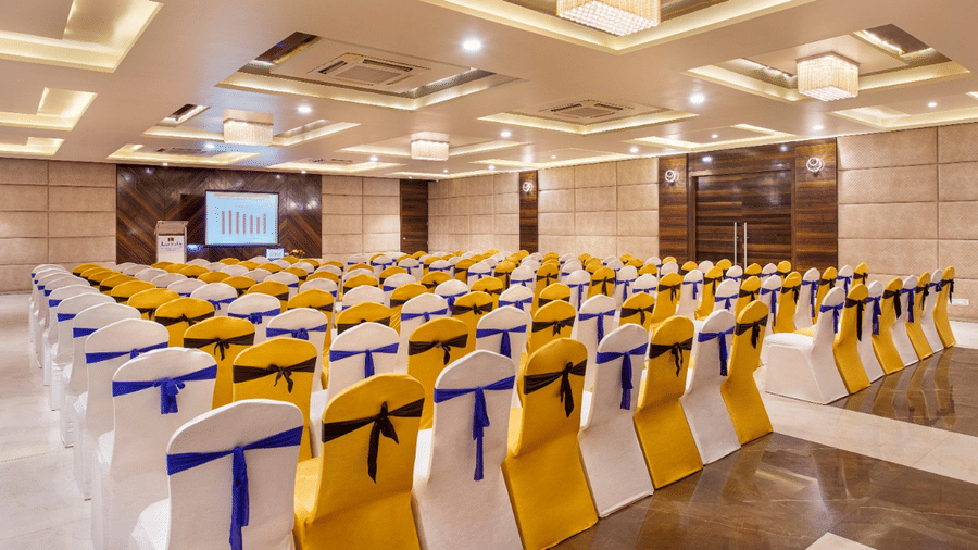 A large banquet hall at Lords Inn Porbandar, is arranged with rows of chairs covered in white and yellow fabric for an event.