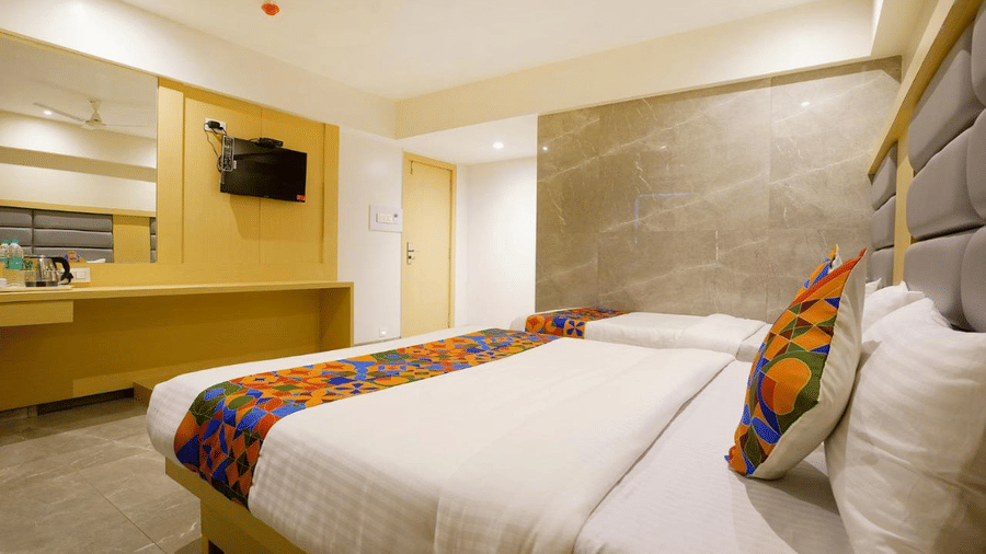 An elegant well furnished room with a twin size bed and a single bed, colourful cushions and runners, wooden furniture with a wall mirror and a mounted T.V. at Shyanu Lords Eco Inn Airoli.