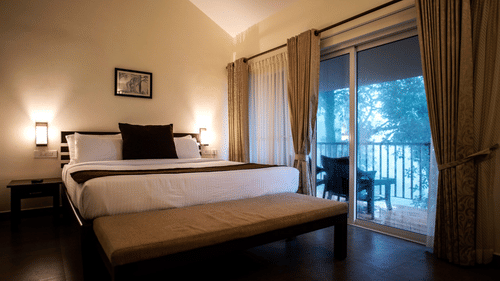 A bedroom features a large bed and sliding glass doors that open to a balcony, providing natural light. - Abad Brookside Lakkidi, Wayanad