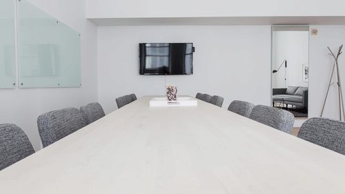 Minimalist modern conference room with a long, light-surfaced table, several textured upholstered chairs, a mounted television, and a glass whiteboard.
