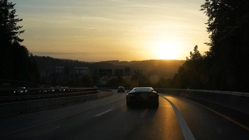 A car driving on a highway during sunset, with the sun low on the horizon and trees on both sides.