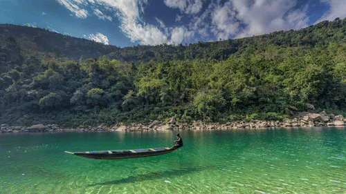 Clear emerald-green river with a small wooden boat floating near a lush, forested hillside under a partly cloudy sky.