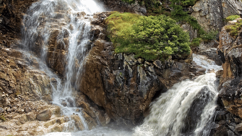 A waterfall cascading over dark rocks and green vegetation into a foaming pool at the bottom.