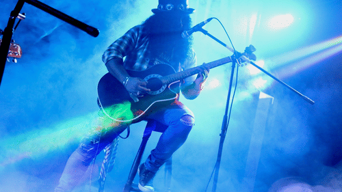 Slash, silhouetted in a top hat and sunglasses, energetically plays guitar on a smoky, brightly lit stage.