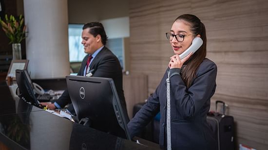 A hotel reception area with two staff members assisting guests, one on the phone and the other at the computer