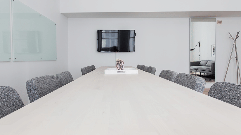 Minimalist modern conference room with a long, light-surfaced table, several textured upholstered chairs, a mounted television, and a glass whiteboard.