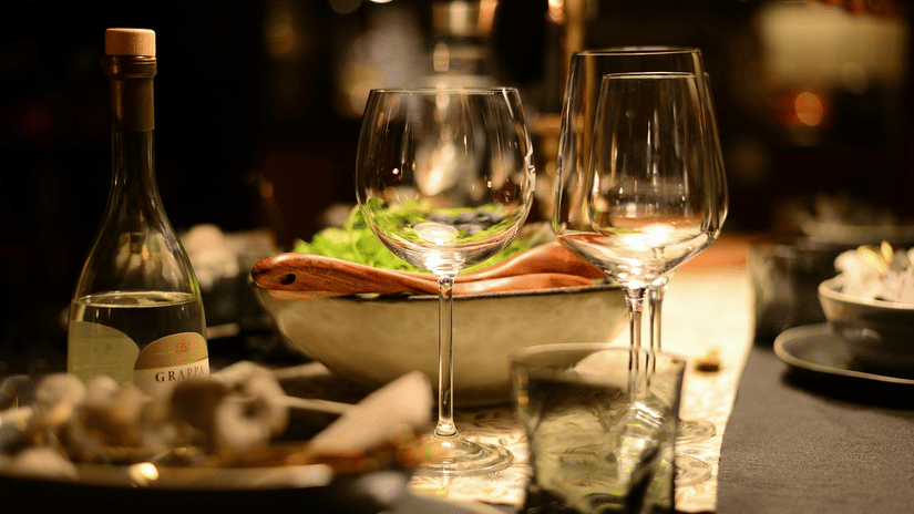 A close up of a dimly lit dinner setting with 2 wine glasses, a bottle, and a plate of food on a black table