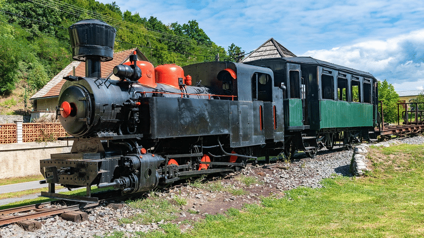 A toy train standing on railway tracks near grassy ground and houses under a bright sky.