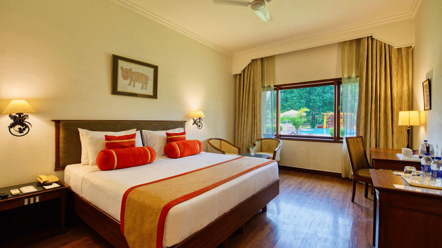 An Deluxe King Pool View bedroom at Clarks Khajuraho with a neatly made bed, bedside lamps, wall artwork, and a window with curtains allowing daylight.