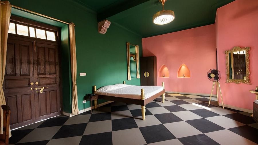 A guest room at Tijara Fort-Palace - 19th Century, Alwar features a bed, a mirror, a standing fan, a door with curtains, and checkered flooring.