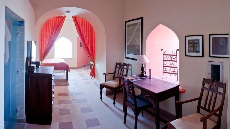 Room with bed visible through arched doorway, curtains, furniture including desk, chairs, and cabinet, framed artwork, and patterned floor tiles - Tijara Fort-Palace - 19th Century, Alwar.