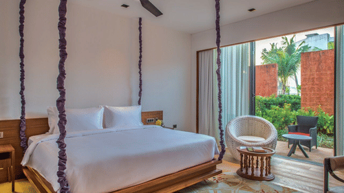 The Deluxe Patio King Room with a large wooden bed hanging from the ceiling on purple rope vines, and an armchair at Azaya Beach Resort, Goa.