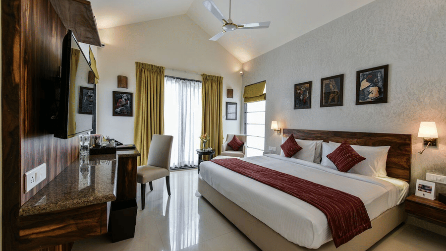 Family room at Tropicana Resort & Spa, Alibaug, featuring a king size bed with white linens and red accents. The room includes vaulted ceilings, a ceiling fan, a wooden headboard, a wall-mounted TV, and a desk area near a large window with gold curtains.