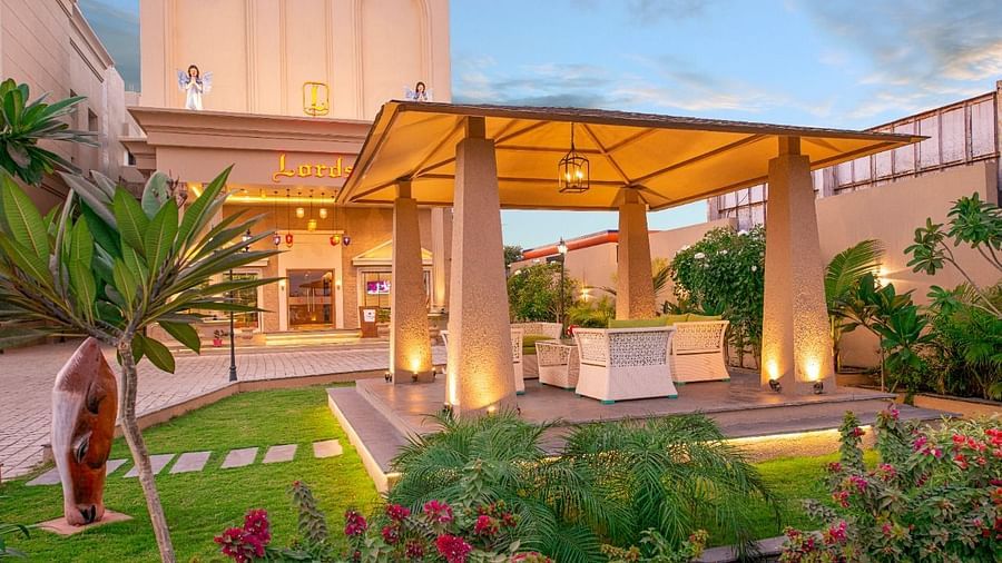 An inviting outdoor area at dusk with a covered seating space supported by pillars, surrounded by well-maintained green lawns, colourful flowering plants, and subtle lighting illuminating the building in the background at Lords Inn Motikhavdi, Jamnagar.