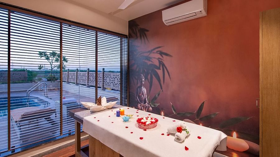 A serene spa treatment room featuring a massage table draped with a white sheet and adorned with red rose petals and decorative items, set against a warm-toned wall with a subtle mural and with natural light coming through large windows overlooking an outd