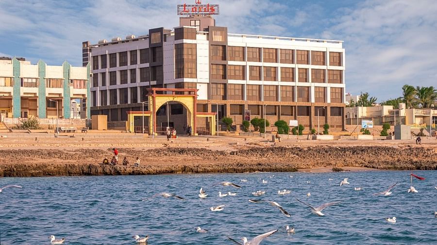 A tan-colored hotel building with multiple floors is situated by the water, with boats visible on the blue sea and a clear sky above.