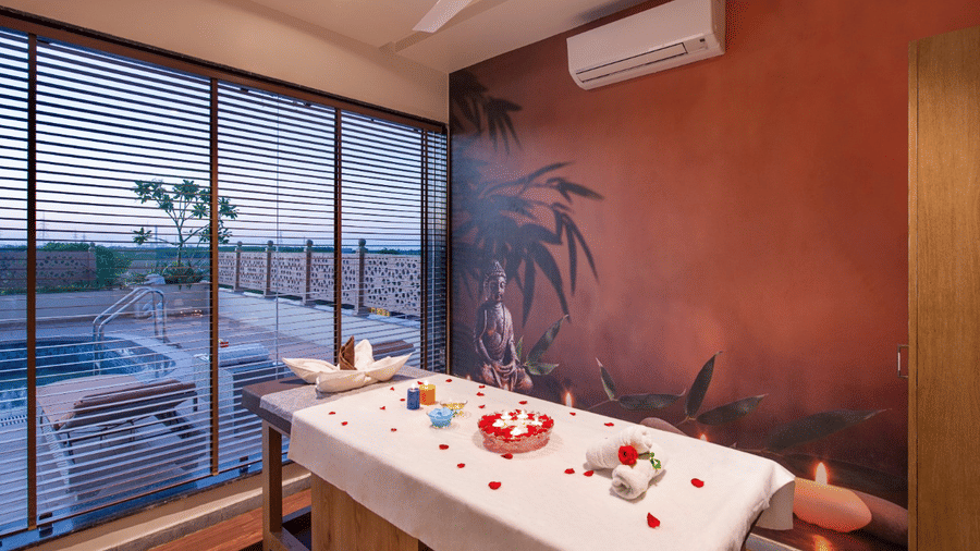 A spa room at Lords Inn Jamnagar featuring a massage table draped with a white sheet and adorned with red rose petals with natural light coming through large windows offering an outdoor view