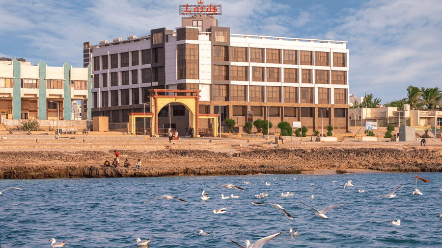 A tan-colored hotel building with multiple floors is situated by the water, with boats visible on the blue sea and a clear sky above.