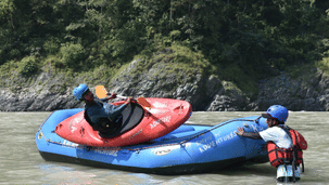 2 people in blue helmets with a red kayak on top of a blue inflatable raft in a river.