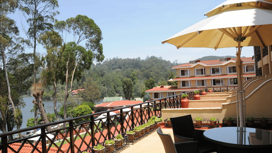 View of the terrace at The Carlton Kodaikanal with outdoor seating, sun umbrella, wooden railing, and a serene lake and hillside beyond.