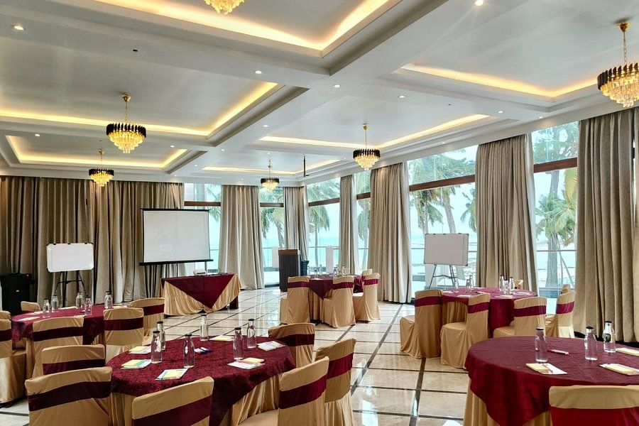 alt-text A large event space with neatly arranged round tables covered in beige and burgundy, illuminated by chandeliers, and large windows offering a view of palm trees outside.